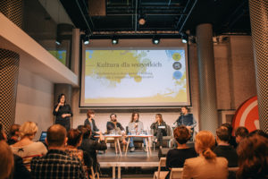 KonfeNa scenie siedzi pięcioro prelegentów. Po lewej stoi tłumaczka języka migowego. Sala pełna widzów.