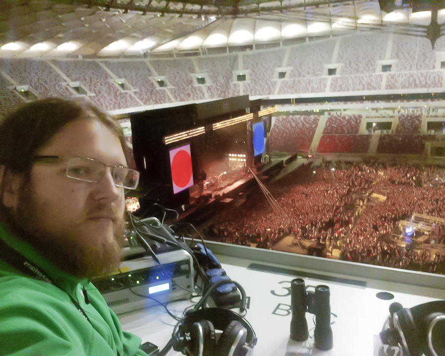 Na pierwszym planie po lewej audiodeskryptor. W tle płyta boiska na Stadionie Narodowym w Warszawie. Na płycie rozstawiona scena. Wokół sceny tłum słucha koncertu.