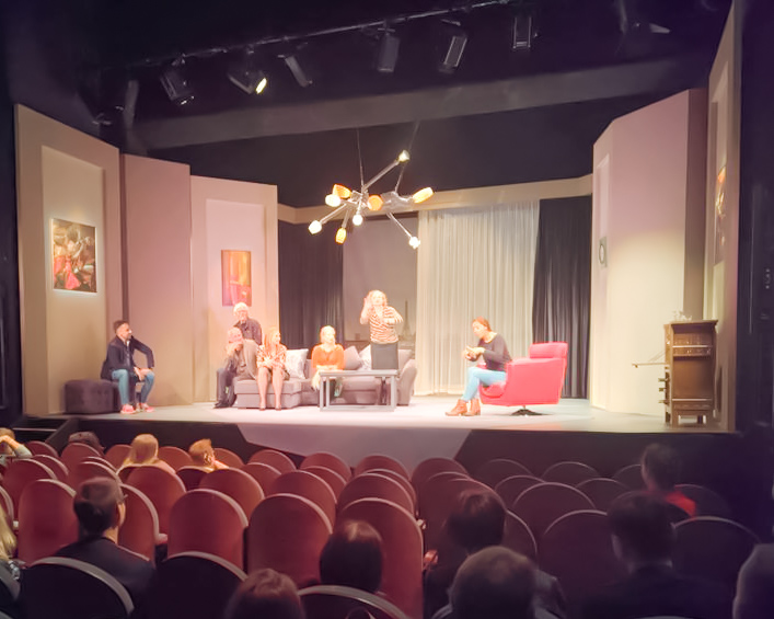 Na scenie teatralnej piątka aktorów siedzi na kanapie. Po prawej, na fotelu, tłumaczka języka migowego.