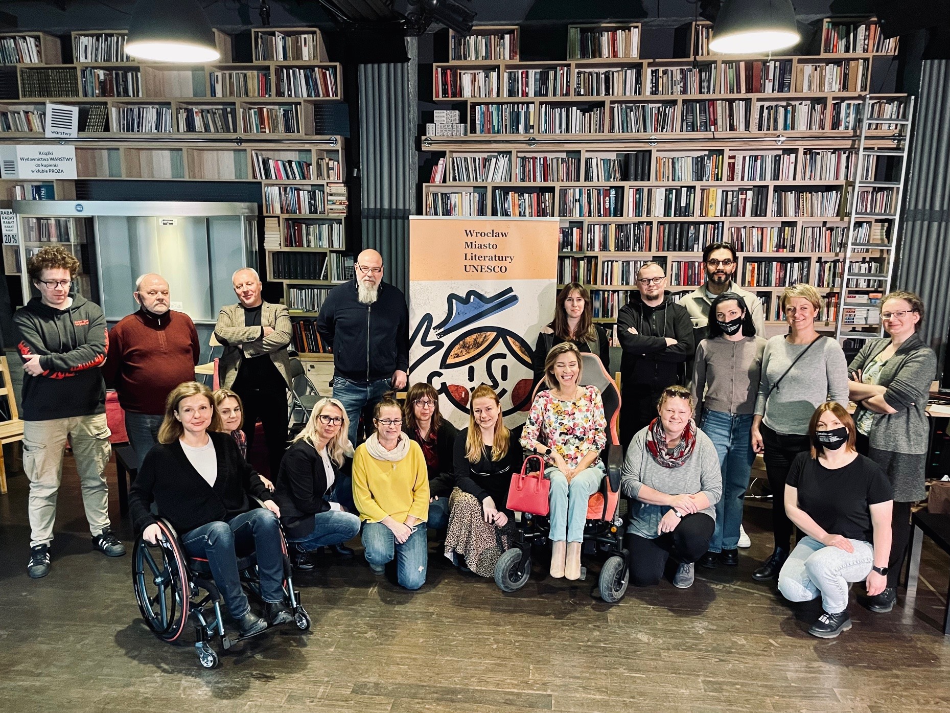 Na tle gigantycznej półki z książkami stoją uczestnicy szkolenia - to kilkunastu pracowniców Wrocławskiego Domu Literatury. Jedna z uczestniczek siedzi na wózku, podobnie jak trenerka Agata Roczniak. Wśród grupy znajdują się też Magda Orłoś i Magda Brumirska z Katarynki.