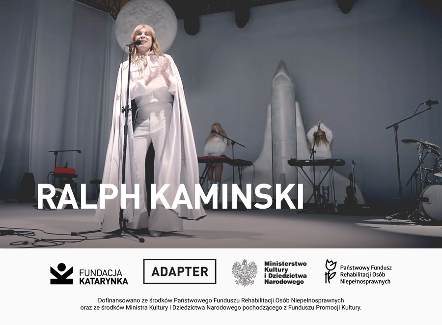 Kolorowe zdjęcie. Scena. Ralf Kaminski w długim płaszczu stoi przy mikrofonie. Śpiewa. Nad głową Ralfa unosi się element dekoracji - księżyc w pełni. W tle dwójka muzyków. Stoją przy kejbordach. Pomiędzy nimi stoi duża kosmiczna rakieta. Poniżej biały pasek. Na nim logotypy: Fundacja Katarynka, Adapter, Ministerstwo Kultury i Dziedzictwa Narodowego, Państwowy Fundusz Rehabilitacji Osób Niepełnosprawnych. Poniżej napis: Dofinansowano ze środków Państwowego Funduszu Rehabilitacji Osób Niepełnosprawnych oraz ze środków Ministra Kultury i Dziedzictwa Narodowego pochodzącego z Funduszu Promocji Kultury.
