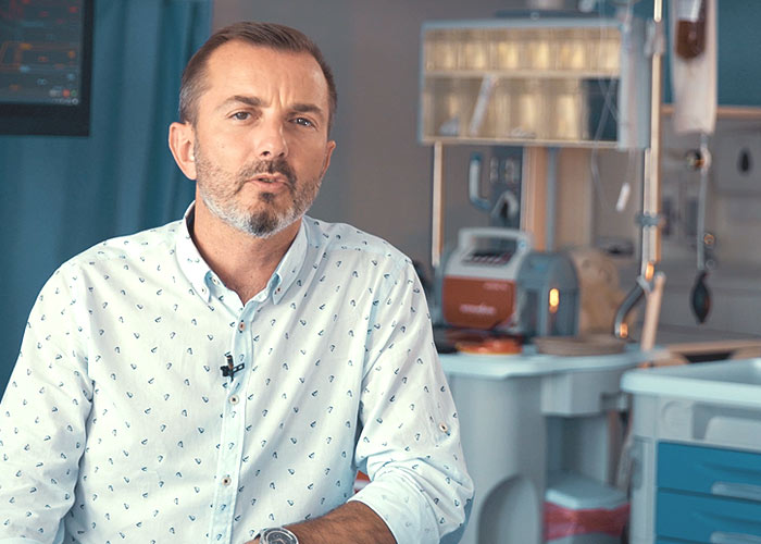 Kolorowe zdjęcie. Po lewej Tomasz Rożek patrzy w obiektyw i opowiada. Ma na sobie białą koszulę we wzorki. Po prawej stoi sprzęt laboratoryjny.