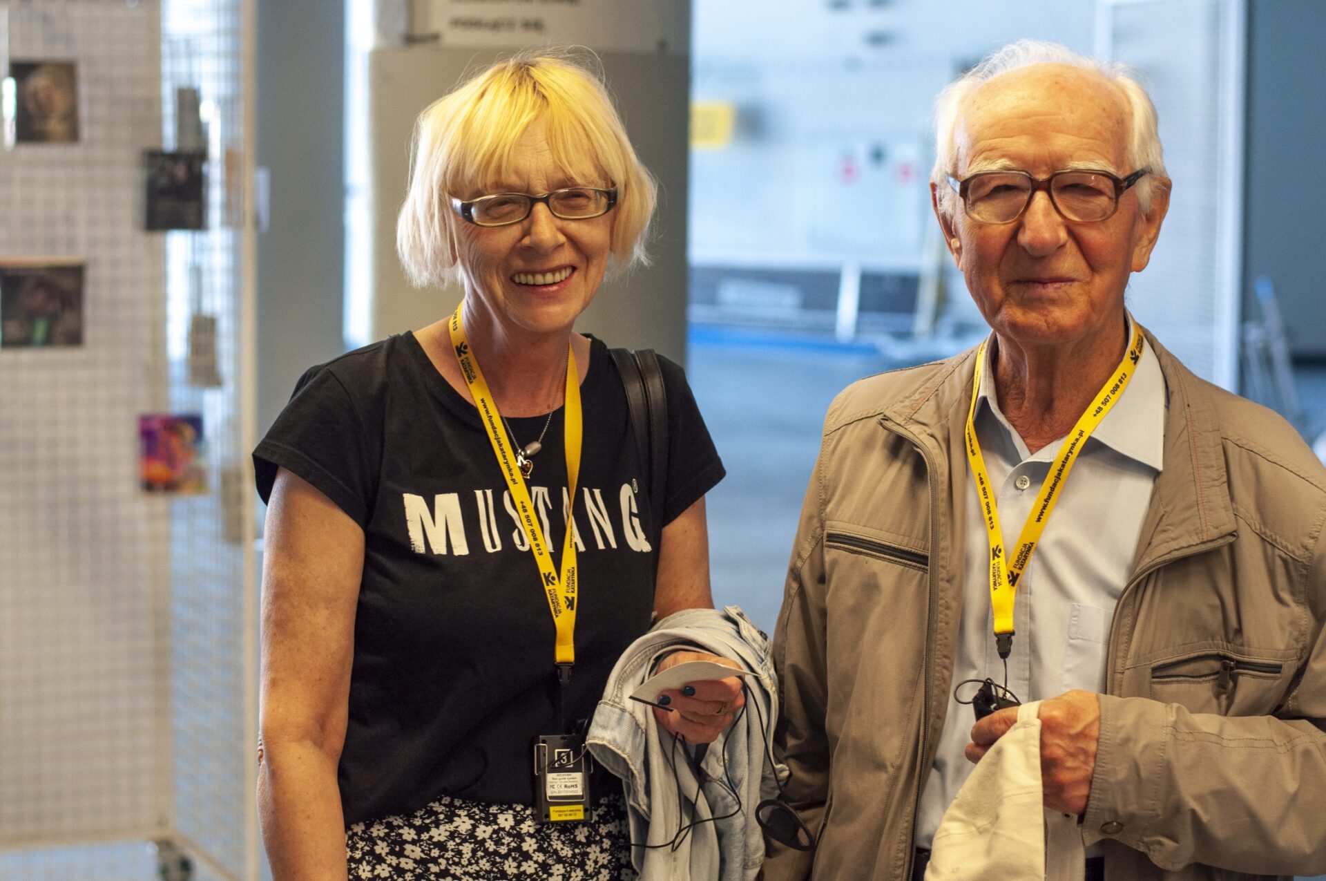 Dwoje seniorów w kinie nowe horyzonty. Po lewej kobieta z krótkimi jasnymi włosami. Po prawej mężczyzna z siwymi włosami. Oboje noszą okulary, uśmiechają się do obiektywu. Na szyjach mają zawieszone na żółtych smyczach odbiorniki do audiodeskrypcji.