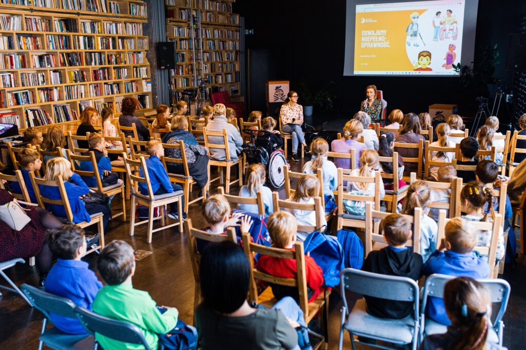 Klub Proza. Pod sceną siedzą Justyna Mańkowska i Agata Roczniak. Przed nimi na krzesłach ustawionych w rzędy siedzą dzieci z wrocławskich szkół. Jedna z dziewczynek siedzi na wózku.