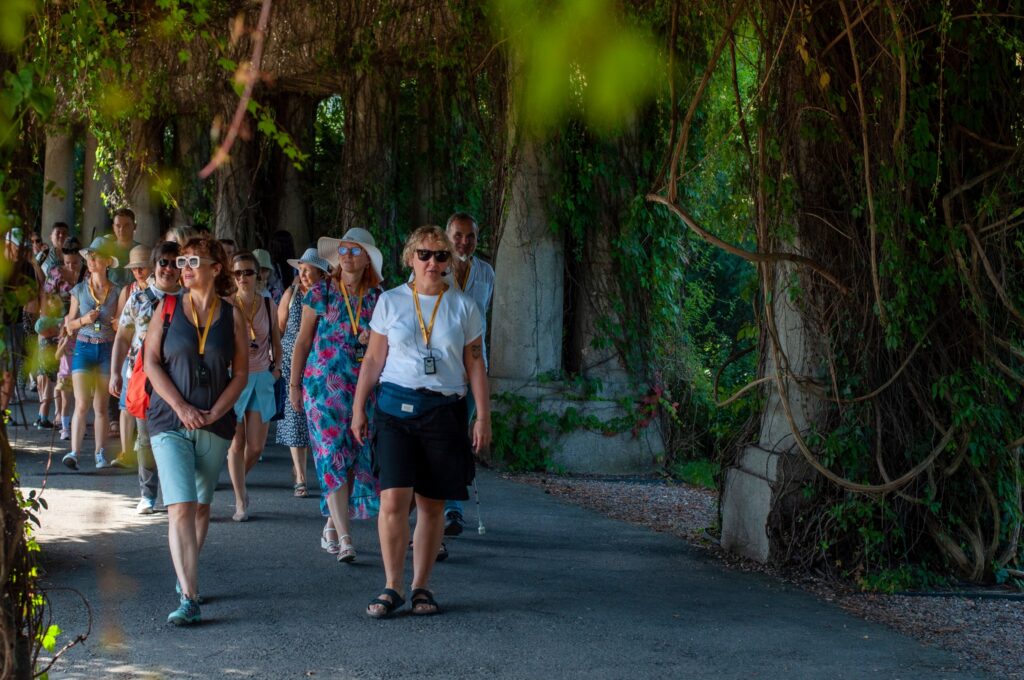 Kolorowe zdjęcie. Park. Ewa Pluta idzie parkową ścieżką. Za nią podąża grupa spacerowiczy. Dookoła wiele drzew.