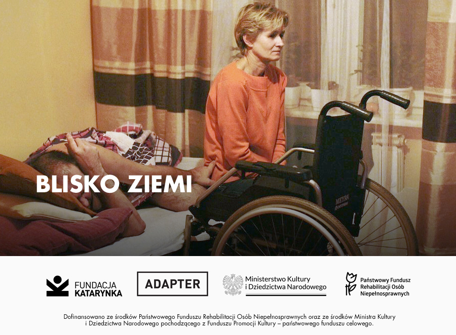 Kolorowy kadr z filmu Blisko ziemi. Kobieta siedzi na brzegu łóżka i patrzy przed siebie. Obok w łóżku leży mężczyzna, a obok łóżka stoi wózek aktywny dla osób z niepełnosprawnością ruchową. Poniże biały pasek, na nim logotypy: Fundacja Katarynka, Adapter, Ministerstwo Kultury i Dziedzictwa Narodowego, Państwowy Fundusz Rehabilitacji Osób Niepełnosprawnych. Dofinansowano ze środków Państwowego Funduszu Rehabilitacji Osób Niepełnosprawnych oraz ze środków Ministra Kultury i Dziedzictwa Narodowego pochodzącego z Funduszu Promocji Kultury - państwowego funduszu celowego.