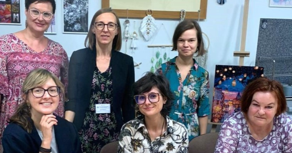 Kolorowe zdjęcie. Sala w Centrum Zdrowia Psychicznego na Ostrowskiego. Zdjęcie grupowe. Przy stoliku siedzą Justyna Mańkowska, Alicja Regiewicz, asystentka zdrowienia. Za nimi stoją dwie terapeutki zajęciowe i asystentka zdrowienia.