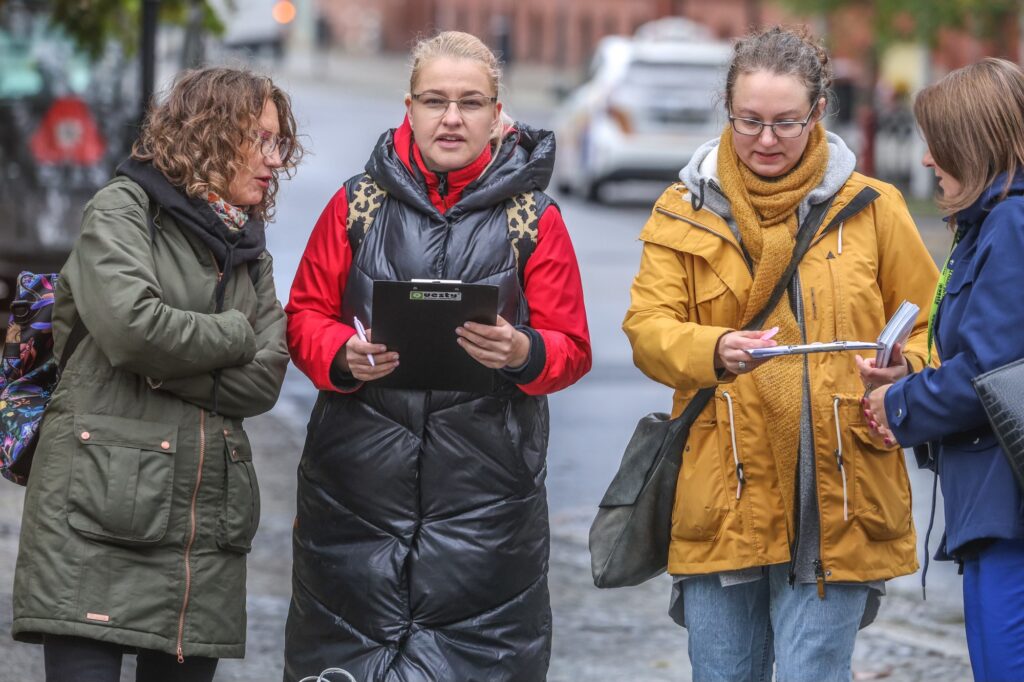 Kolorowa fotografia. Od lewej stoi Ania Brzegowa Jakbczyk, Ola Szalek Palmowska i Magda Brumirska Zielińska. Ola trzyma w ręce podkładkę z ulotką gry.