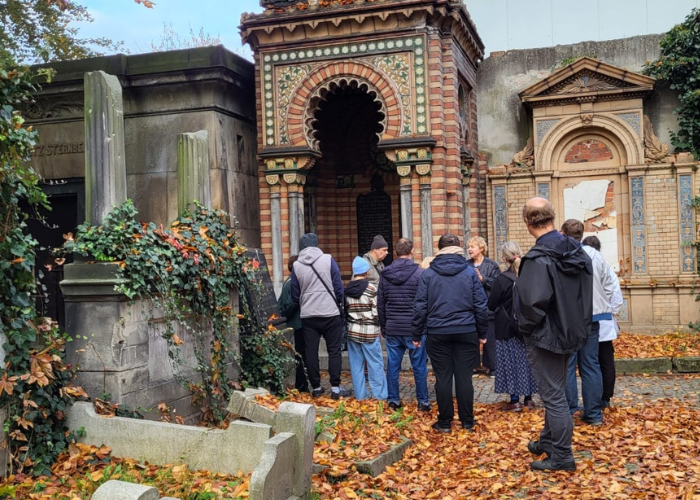 Kolorowe zdjęcie. Grupa uczestników Klubu Pacjenta podczas oprowadzania po Starym Cmentarzu Żydowskim we Wrocławiu. Zwiedzający stoją przed zabytkowym, bogato zdobionym mauzoleum, otoczonym jesiennymi liśćmi i porośniętymi bluszczem nagrobkami. Słuchają przewodniczki Ewy Pluty.