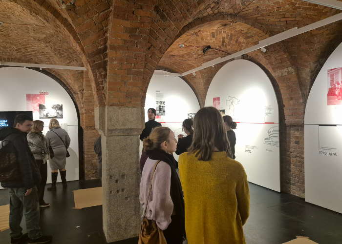 Kolorowe zdjęcie. Grupa zwiedzających z Klubu Pacjenta przewodnika na wystawie w Krupa Art Galleri. Są toczeni ceglaną architekturą i tablicami informacyjnymi o twórczości artysty Fangora.