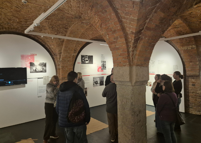 Kolorowe zdjęcie. Grupa zwiedzających z Klubu Pacjenta przewodnika na wystawie w Krupa Art Galleri. Są toczeni ceglaną architekturą i tablicami informacyjnymi o twórczości artysty Fangora.