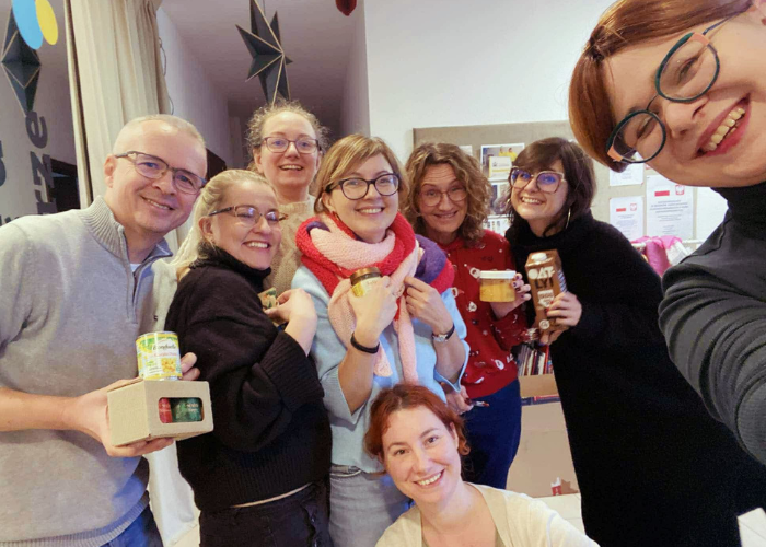 Kolorowe zdjęcie w Pracowni Dostępnej Kultury. Ekipa kataryniarzy uśmiecha się i patrzy w obiektyw. Od lewej Mariusz, Ola, Magda, Justyna, Ania, Kasia, Ala i Daniela. Trzymają różne przedmioty, które wygrali w kole fortuny między innymi czekoladowe mleko owsiane, miody czy kosmetyki.