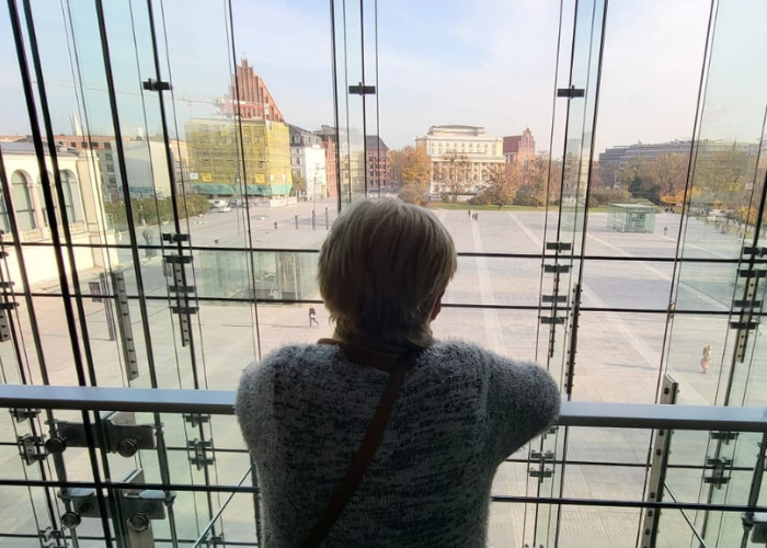 Kolorowe zdjęcie. Jedna z odbiorczyń Klubu Pacjenta w Narodowym Forum Muzyki stoi przy szklanej balustradzie i patrzy na przestronny plac z panoramą miasta Wrocław w tle.