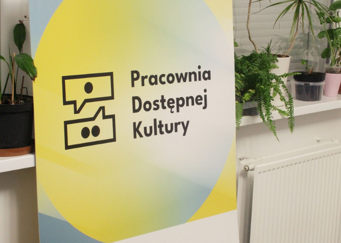 Kolorowe zdjęcie. Żółty niebieski baner z napisem Pracownia Dostępnej Kultury stoi oparty i parapet. Na parapecie rośliny doniczkowe.