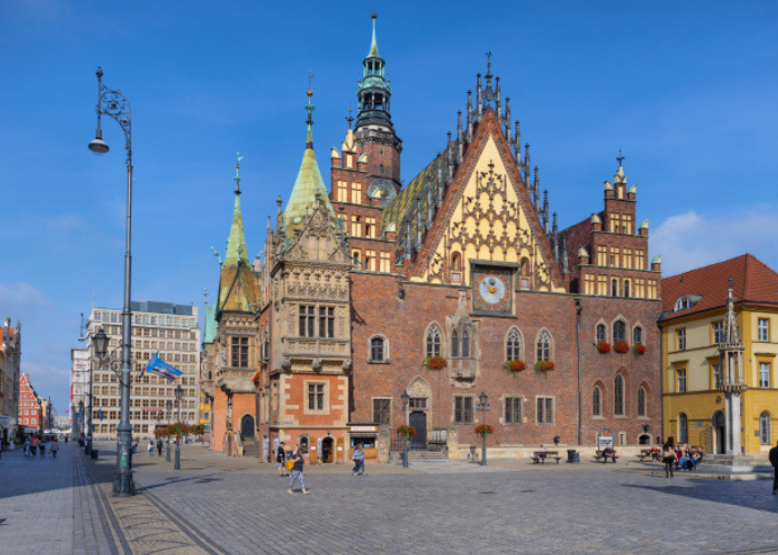 Kolorowe zdjęcie. Gotycki Ratusz we Wrocławiu na Rynku. Budynek wyróżnia się bogato zdobioną fasadą, zegarem astronomicznym i charakterystycznym dachem z wieżyczkami. Na placu przed ratuszem spacerują ludzie.