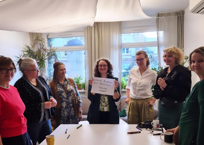 Kolorowe zdjęcie grupowe w Pracowni Dostępnej Kultury. Koordynatorki dostępności stoją przy białym prostokątnym stole. Magda Brumirska trzyma kartkę z napisem Grupa Kultura Dla Wszystkich.