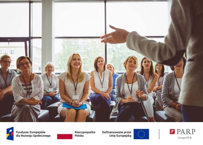Kolorowe zdjęcie. Grupa kobiet uczestniczy w prezentacji, siedząc w jasnej sali konferencyjnej. Uśmiechają się i słuchają prelegenta, trzymając notesy i długopisy. Na dole zdjęcia znajdują się logotypy: Fundusze Europejskie dla Rozwoju Społecznego, Rzeczpospolita Polska, Unia Europejska oraz PARP Grupa PFR.