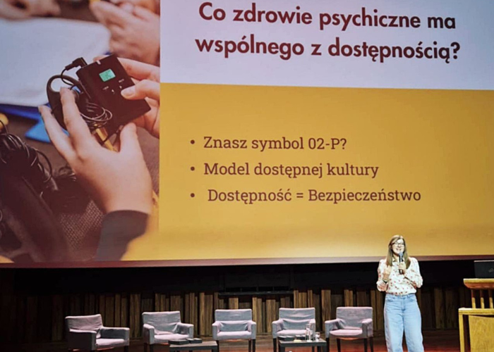 Kolorowa fotografia. Sala Wielka w Centrum Kultury Zamek w Poznaniu. Justyna Mańkowska Kaczmarek stoi na scenie i mówi do mikrofonu. Za sobą ma ekran rzutnika. Na slajdzie napis "Co zdrowie psychiczne ma wspólnego z dostępnością?