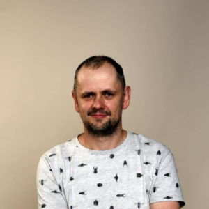 zdjęcie uśmiechniętego mężczyzny – to audiodeskryptor Bartek Wokan, ubrany w jasną koszulkę, na tle gładkiej, beżowej ściany.