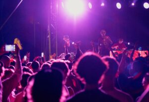 Zdjęcie z koncertu. W fioletowym świetle widać tłum ludzi skierowanych ku scenie. Jedna osoba nagrywa koncert telefonem. Po lewej stronie sceny tłumaczka PJM.