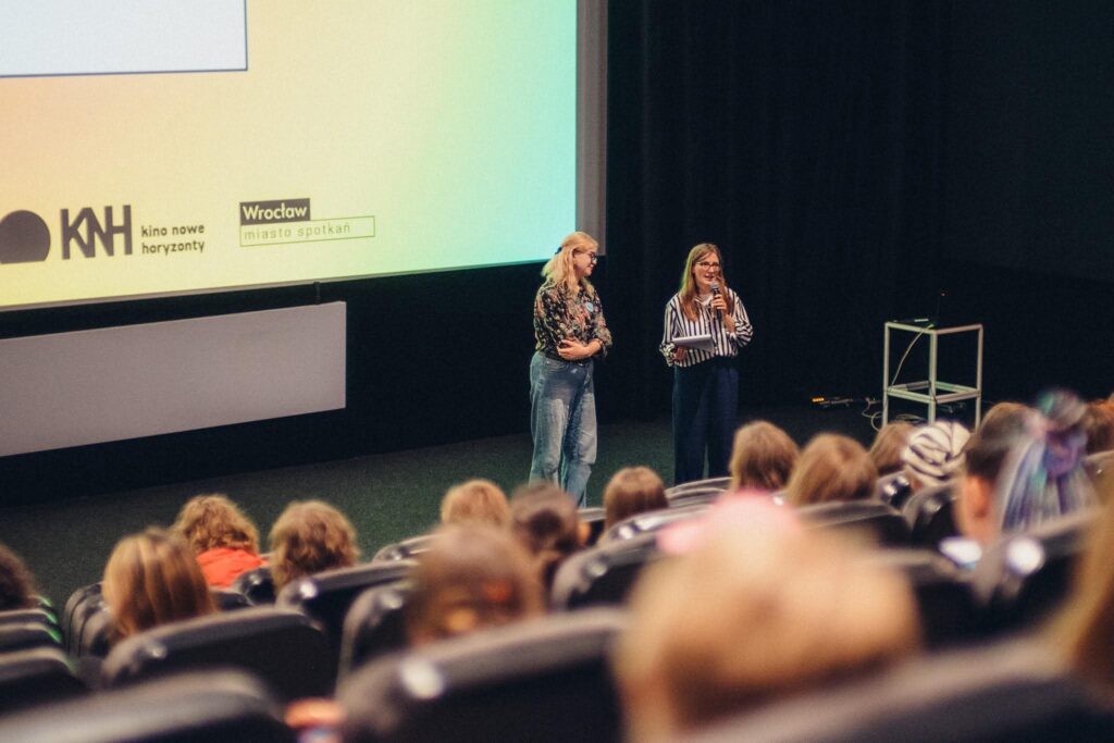 Zdjęcie sceny w kinie. Na środku stoją dwie, młode kobiety. Mają długie, jasne włosy. W ręce trzymają mikrofon. Przed nimi na fotelach siedzi grupa słuchaczy. W tle na ekranie wyświetla się kolorowe tło.