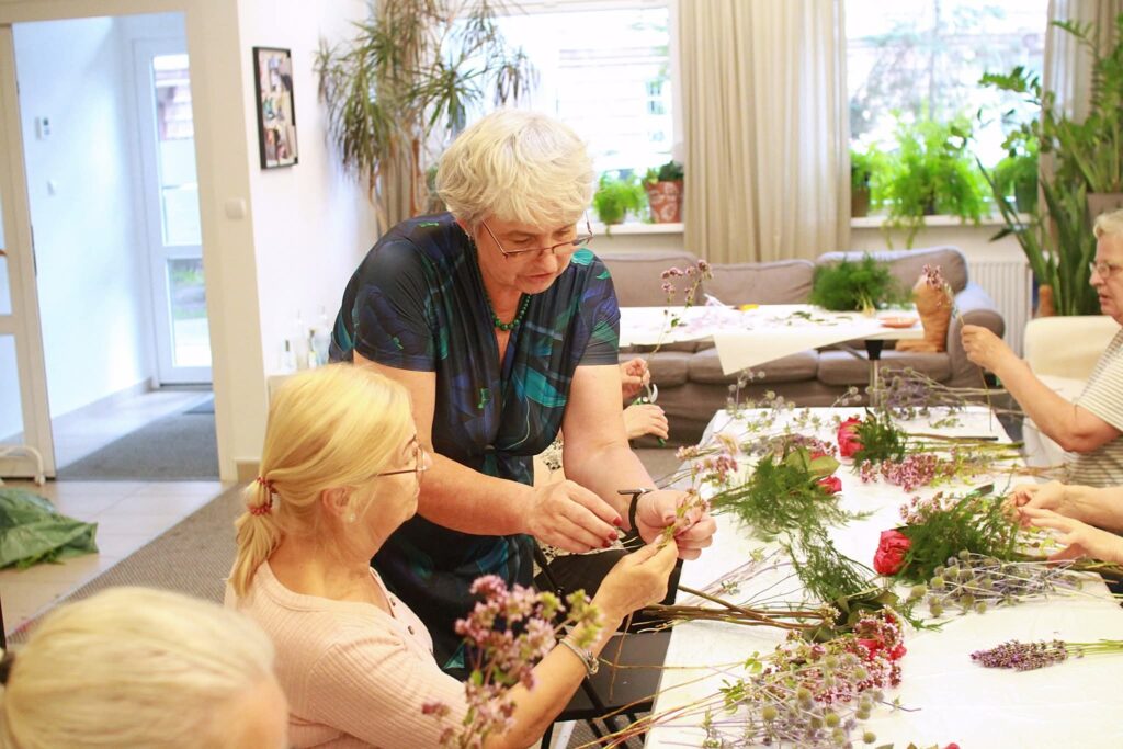 Na zdjęciu dwie seniorki. Wspólnie plotą bukiet, wokół nich na stoliku leżą pozostałe kwiaty i zielone gałązki.