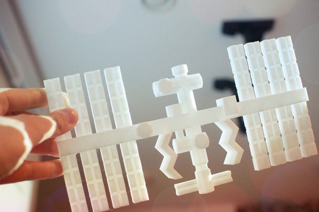 Zdjęcie modelu 3D stacji kosmicznej. Model jest biały. Po lewej i prawej stronie modelu ułożone są dwa prostokąty, imitujące skrzydła, łączą środek modelu stacji.