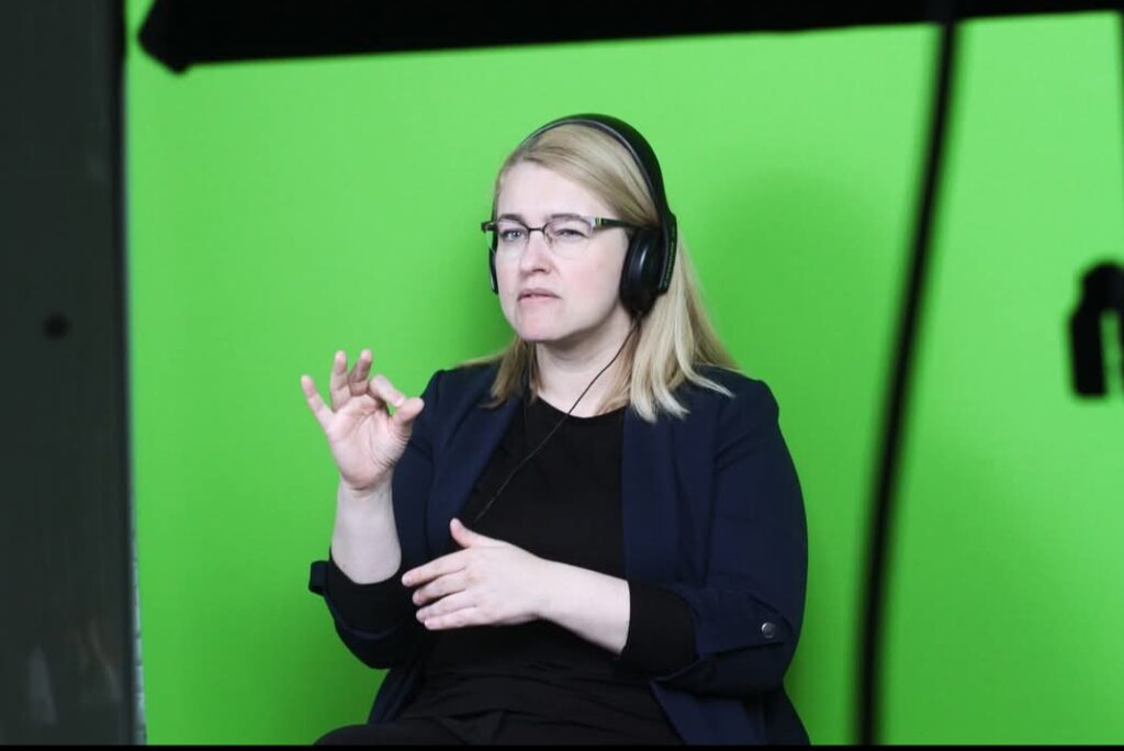 Na zdjęciu kobieta w blond włosach i czarnym ubraniu. Założone ma okulary i duże słuchawki. Wykonuje gesty dłońmi, tłumacząc na PJM. W tle zielone tło.