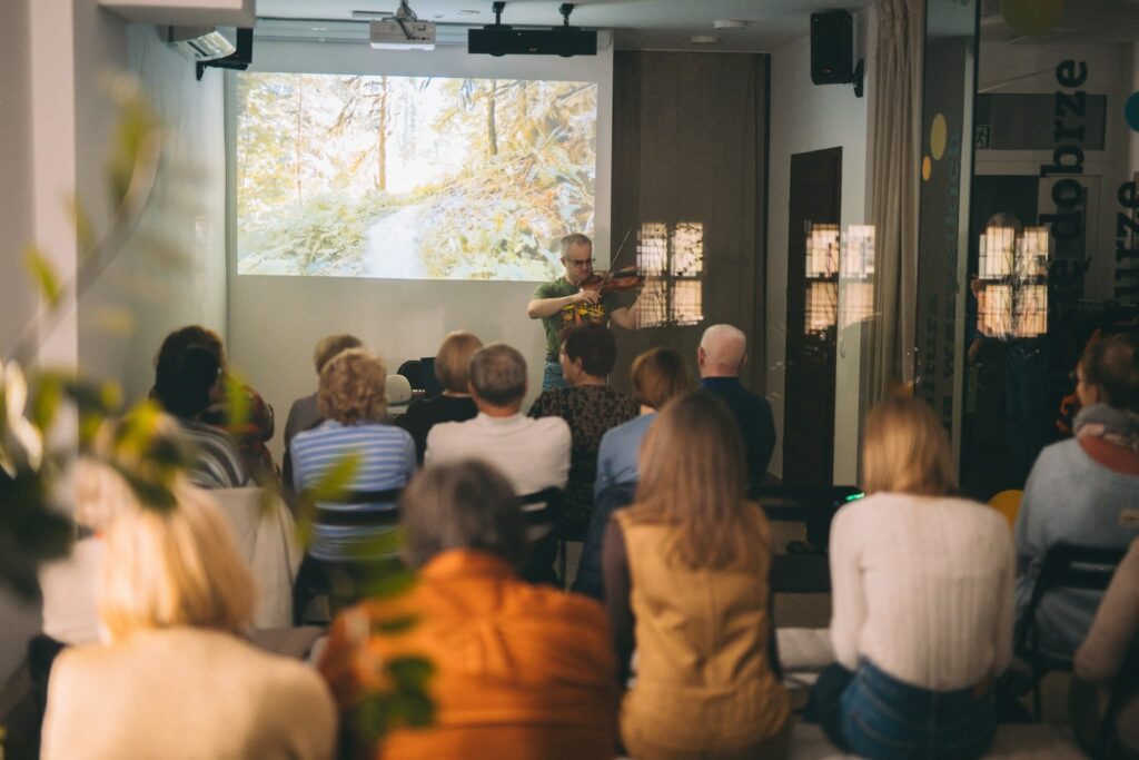 Mężczyzna w Pracowni Dostępnej Kultury gra na skrzypcach. Za nim włączono na rzutniku zdjęcie drzew. W mężczyznę wpatruje się widownia, kilkanaście osób siedzi na krzesłach.