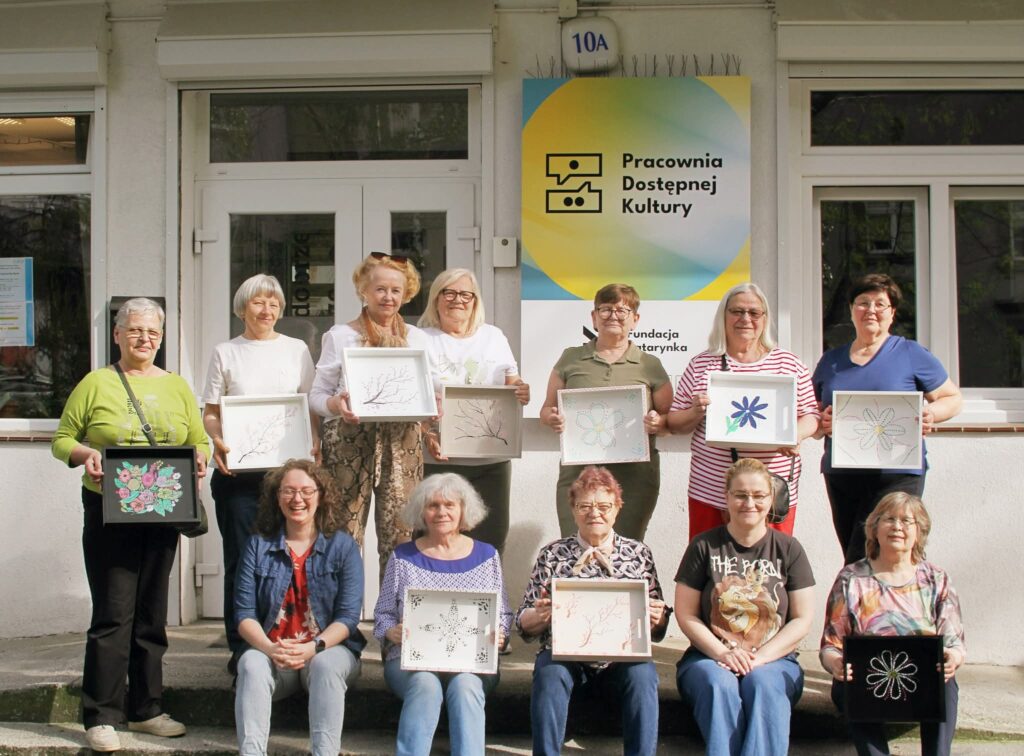 Na zdjęciu jest grupa dwunastu, uśmiechniętych kobiet ustawionych przed budynkiem z szyldem Pracownia Dostępnej Kultury. Każda z kobiet trzyma wykonane prace artystyczne: kolorowe obrazki i hafty umieszczone w ramkach.