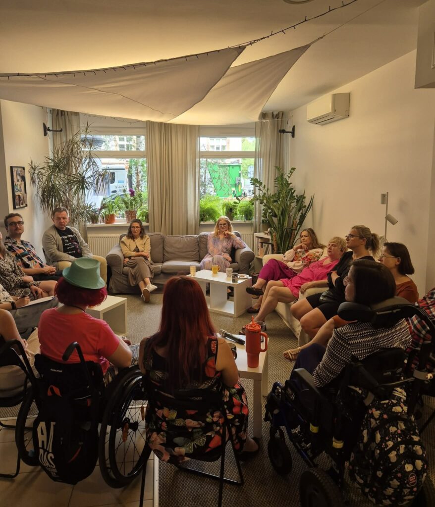 Na zdjęciu grupa osób siedzących w kręgu w jasnym pomieszczeniu. W pomieszczeniu znajdują się sofy, niskie stoliki i dużo roślin w doniczkach. Część osób porusza się na wózkach inwalidzkich.