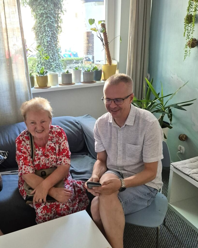 Starsza kobieta w sukience siedzi na kanapie obok mężczyzny, który trzyma telefon. Oboje są uśmiechnięci. W tle okno i dużo roślin.