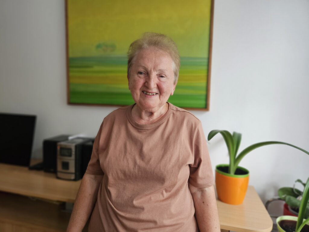 Na zdjęciu starsza kobieta uśmiecha się do aparatu, stojąc w pomieszczeniu. Ubrana jest w jasną koszulkę. W tle duży na jasnej ścianie wisi kolorowy obraz.