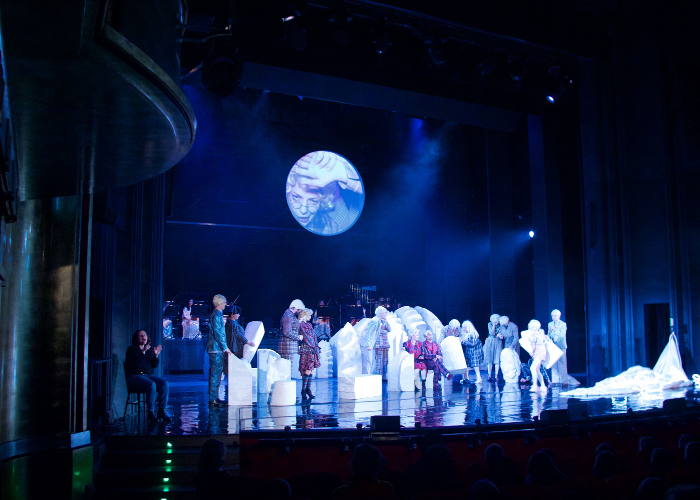 Kolorowe zdjęcie. Scena teatralna ze spektaklu ‘Alicja’. Na scenie stoi grupa aktorów w kolorowych, fantazyjnych kostiumach. W tle widoczna jest orkiestra oraz okrągła projekcja z twarzą kobiety w okularach. Scenografia składa się z dużych, białych elementów przypominających abstrakcyjne kształty. Na scenie po lewej stronie siedzi Ela Resler. Tłumaczy spektakl na polski język migowy.