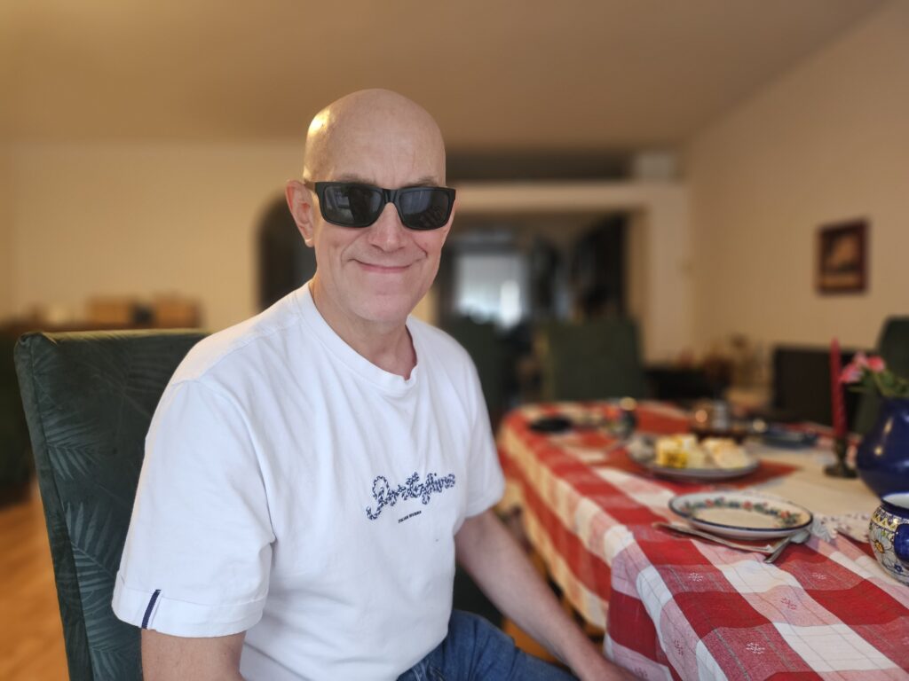 Mężczyzna w jasnej koszulce i w ciemnych okularach uśmiecha się do aparatu, siedząc przy nakrytym stole.