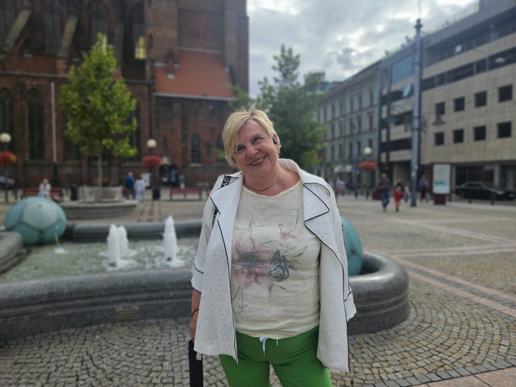 Na zdjęciu uśmiechająca się kobieta w blond włosach. Ma na sobie jasne ubrania. W tle fontanna oraz budynki.
