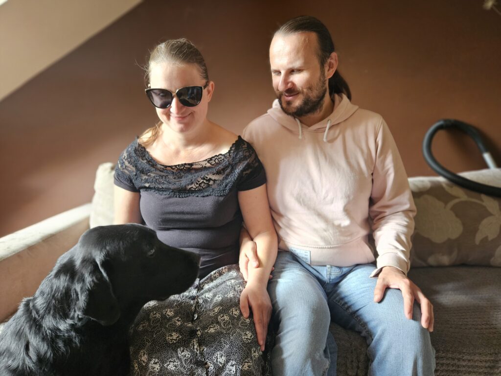 Kobieta w ciemnych okularach i mężczyzna w jasnych ubraniach siedzą na kanapie. Trzymają się za ręce. Przed nimi siedzi czarny pies.