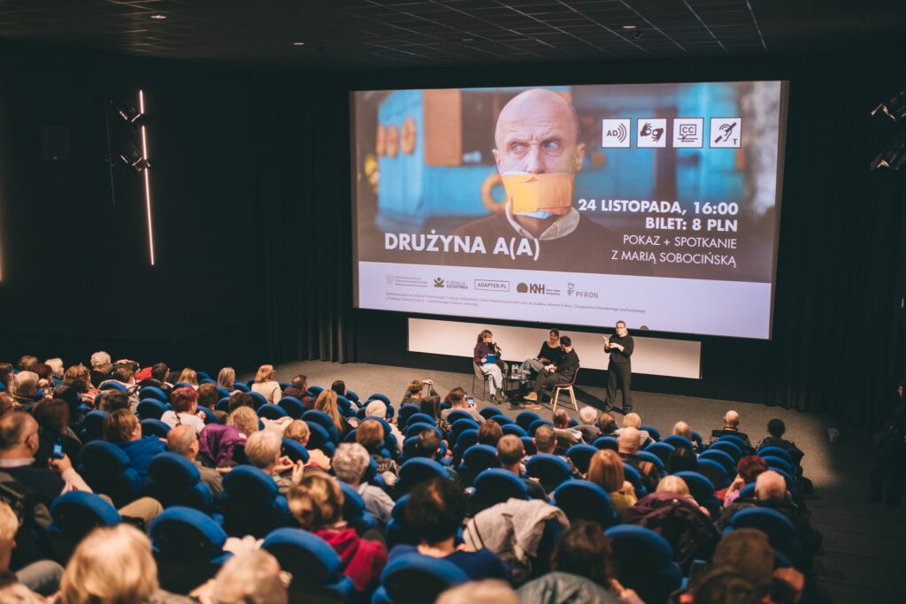 Zdjęcie sali kinowej. Na ekranie wyświetla się plakat promujący film Drużyna A(A). Na scenie kilka osób przemawia do mikrofonów. Przed nią siedzi tłum osób.