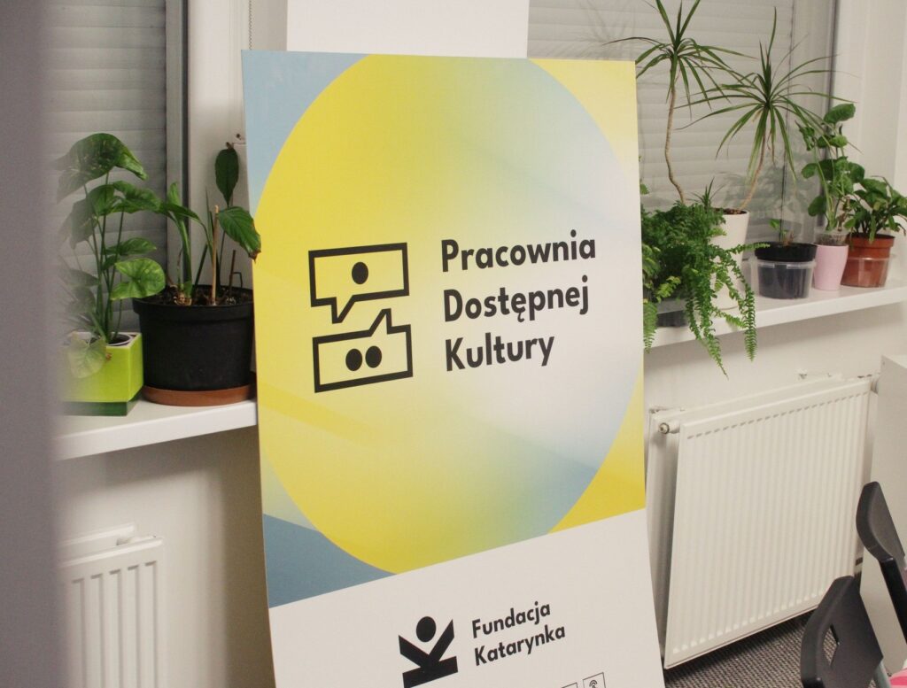 Zdjęcia pomieszczenia. O ścianę stoi oparty duży baner w jasnych kolorach i z napisem Pracownia Dostępnej Kultury, Fundacja Katarynka. Obok niego, na parapecie ustawione są kwiaty w doniczkach.