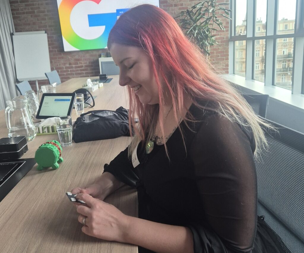 Uśmiechnięta kobieta w czerwonych włosach siedzi przy długim stole. W rękach trzyma mały prototyp 3D. W tle na ścianie wisi logo Google.