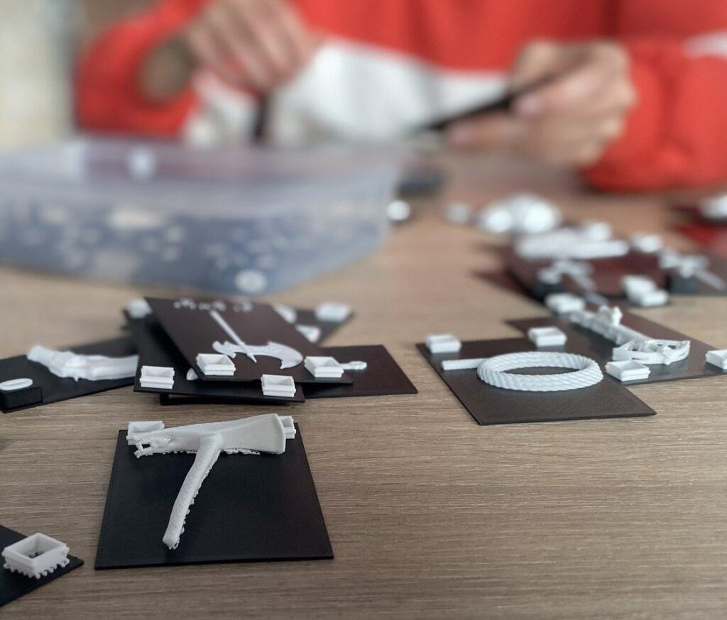 Zdjęcie zestawu wydrukowanych w technologii 3D elementów, które leżą na ciemnych kartach. Elementy mają różne kształty, są białe. W tle zarys osoby przy stole.