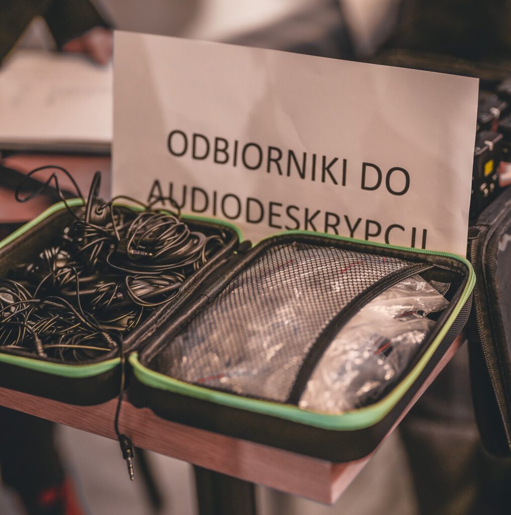 Zdjęcie odbiorników do audiodeskrypcji w futerale. Obok ustawiona kartka z napisem: odbiorniki do audiodeksrypcji.