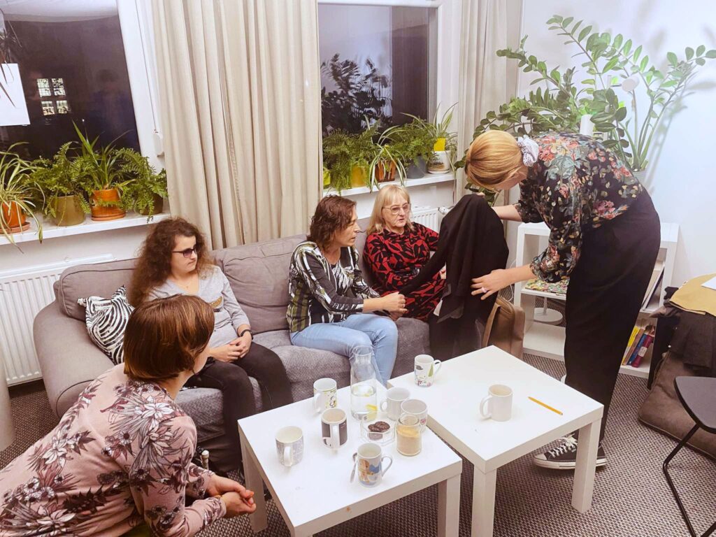 Zdjęcie pomieszczenia. Wokół stolika z kubkami siedzą cztery kobiety. Przed nimi schyla się prowadząca, trzymając w rękach czarną bluzkę. W tle dużo roślin i długie zasłony.