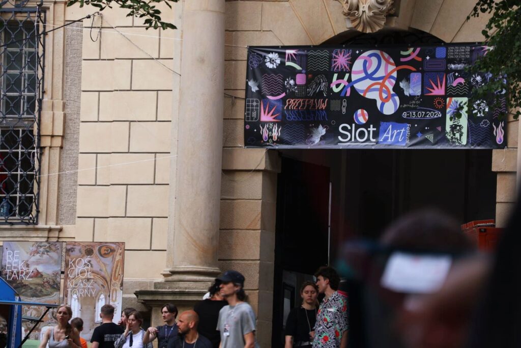 Zdjęcie jasnej ściany budynku z kolumnami. Nad wejściem wisi kolorowy baner "Slot Art". Wokół zgromadzeni są ludzie.
