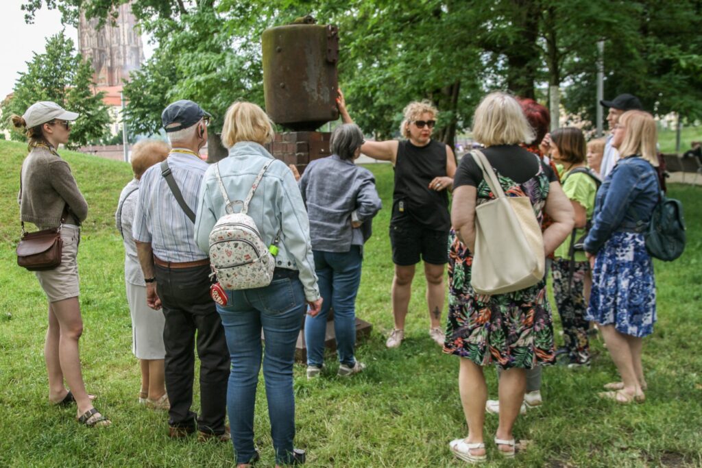 Zdjęcie grupy osób na trawie w parku. Kobieta z odbiornikiem do audiodeksrypcji trzyma rękę na rzeźbie. Wokół drzewa.